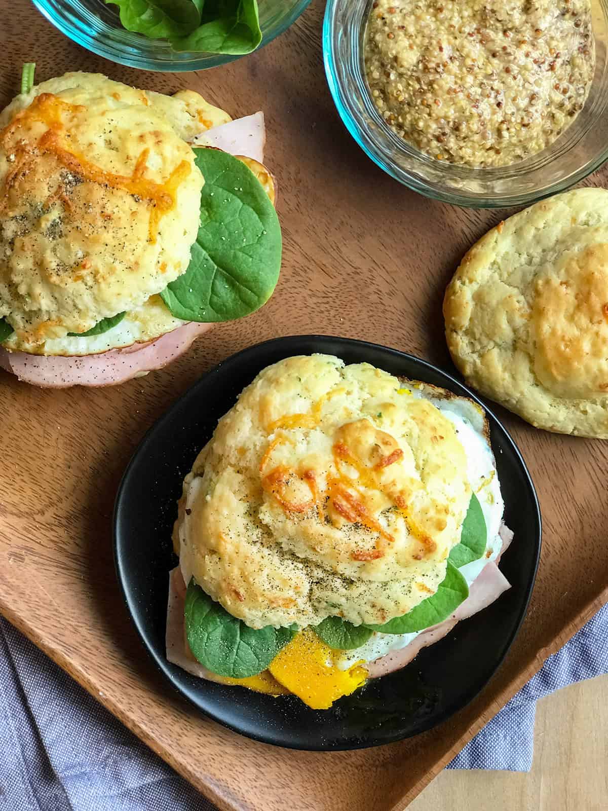 Scottish Breakfast Sandwich biscuits filled with ham, spinach, and a fried egg, served on a plate with a bowl of whole grain mustard and an extra biscuit on a wooden tray.