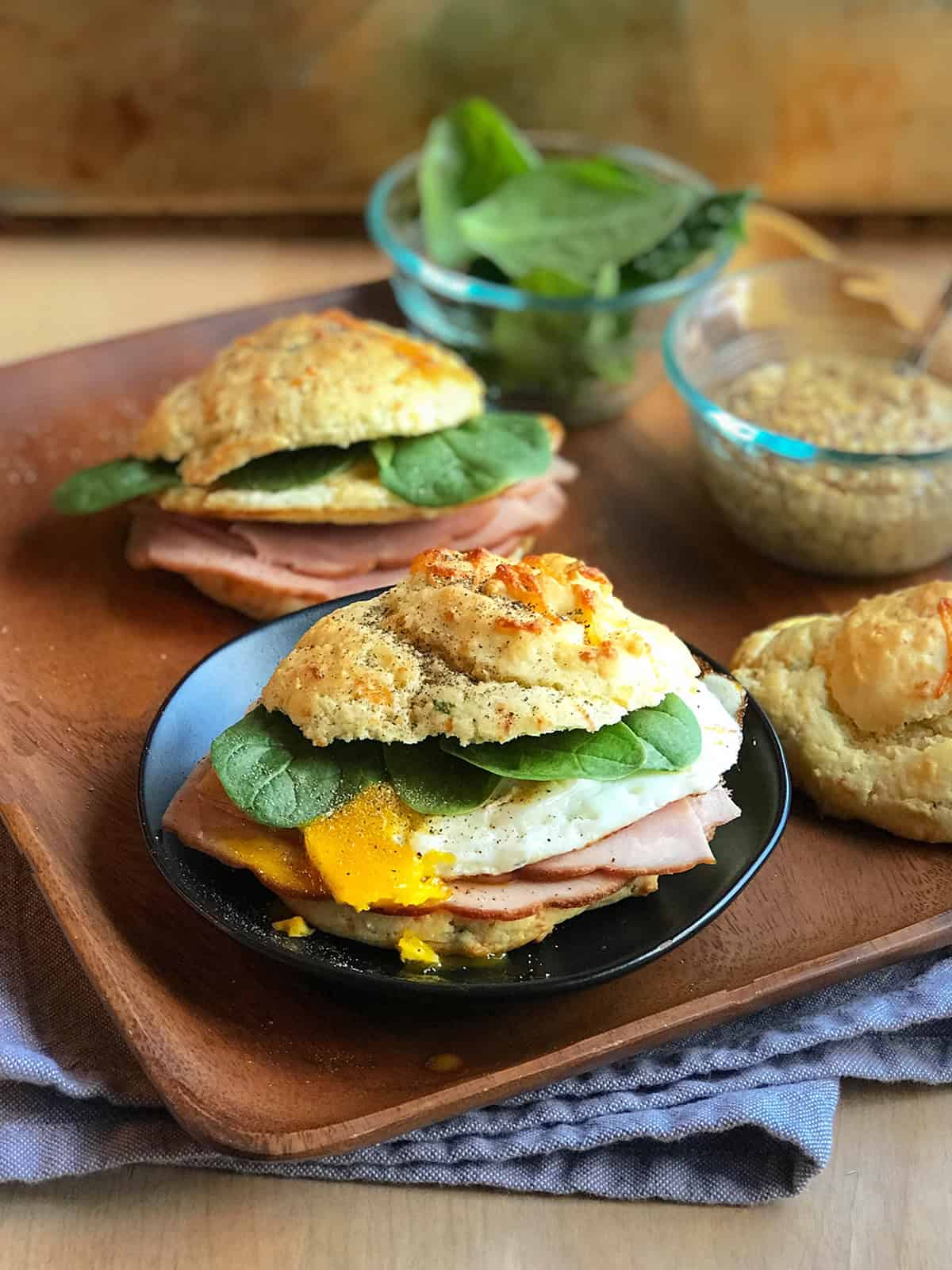 A Scottish Breakfast Sandwich featuring ham, spinach, and a fried egg on a biscuit, with mustard, fresh spinach leaves, and an extra biscuit in the background.