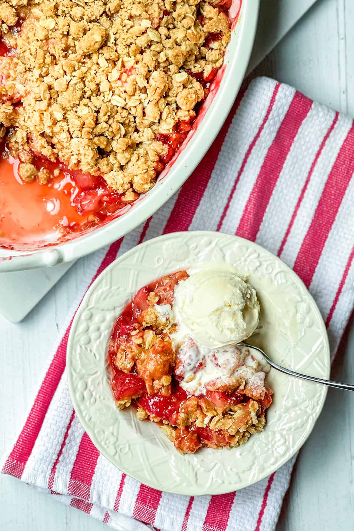 A plate of strawberry crisp with a scoop of vanilla ice cream sits next to a baking dish of crumble, all arranged on a red and white striped cloth.