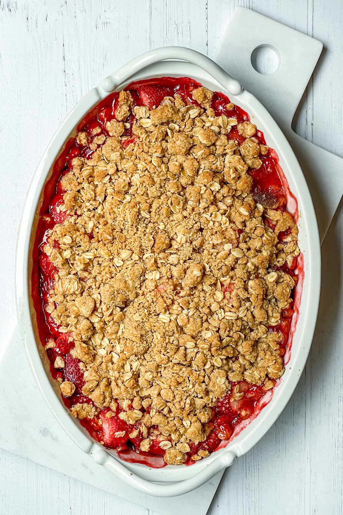 An oval ceramic dish with strawberry crisp, featuring a golden-brown oat and sugar topping, set on a white wood surface.