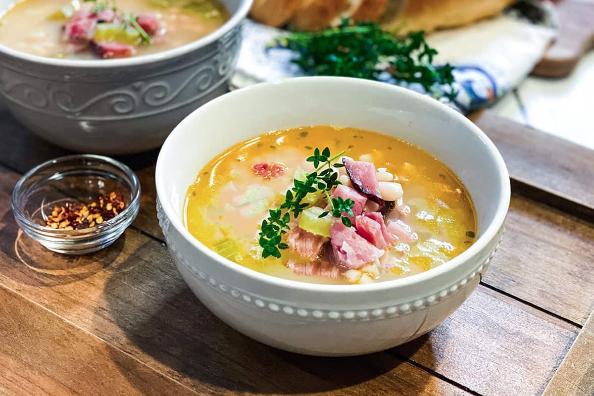 A bowl of ham bone soup and beans garnished with fresh herbs sits on a wooden surface, accompanied by a small bowl of red pepper flakes. Another bowl of hearty soup appears in the background.