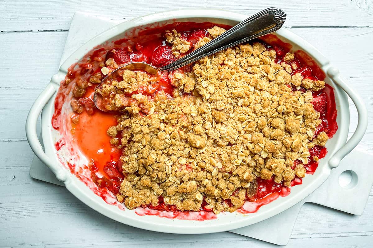 A white oval baking dish filled with strawberry crisp, partially served with a spoon, placed on a white wooden surface.