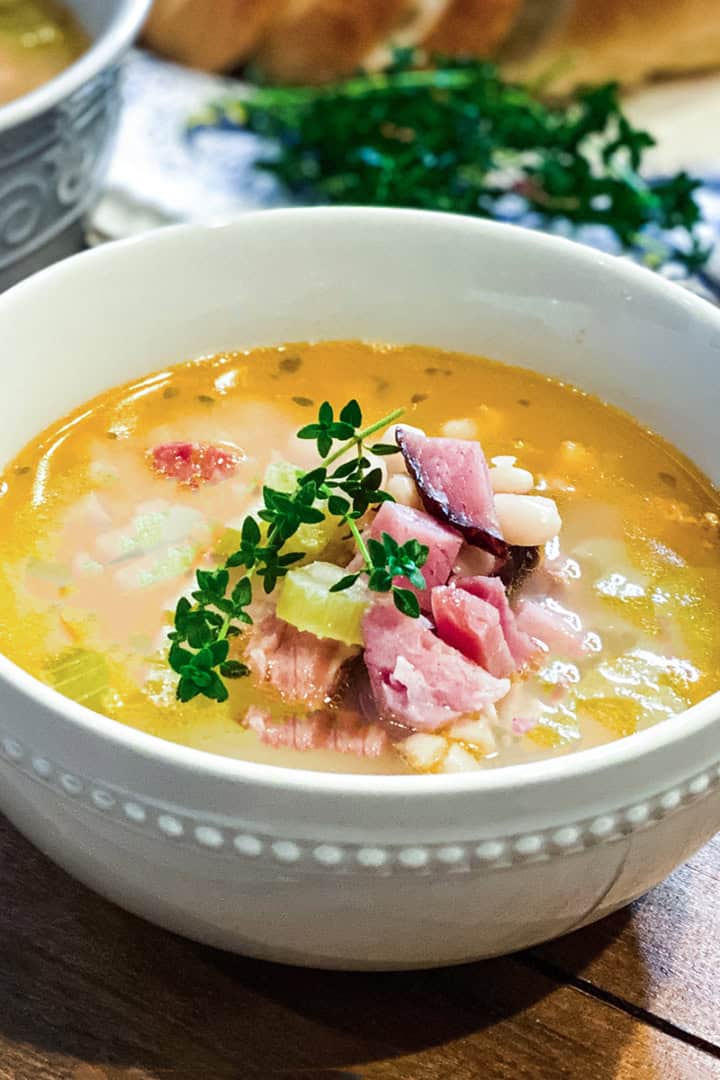 A white bowl of ham bone soup with beans, diced ham, and herbs is garnished with a sprig of fresh thyme and placed on a wooden surface.