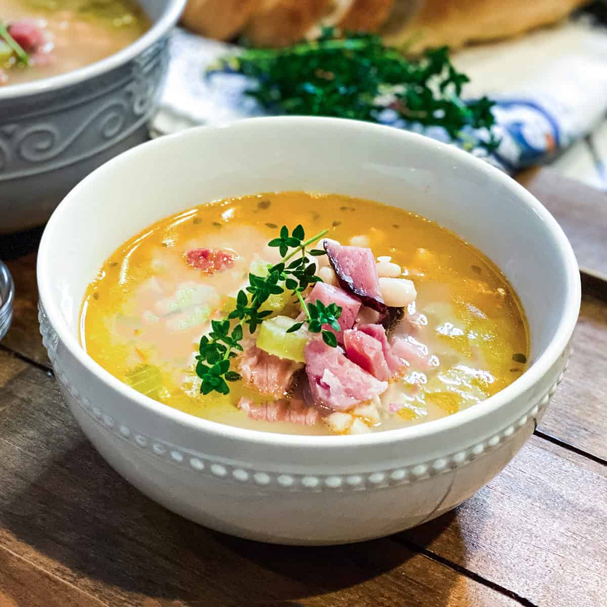 A white bowl of ham bone soup with beans, diced ham, and herbs is garnished with a sprig of fresh thyme and placed on a wooden surface.