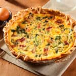 Easy bacon spinach quiche in a glass pie plate resting on a wooden surface with more brown eggs in the background.