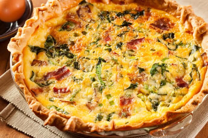 Easy bacon spinach quiche in a glass pie plate resting on a wooden surface with more brown eggs in the background.