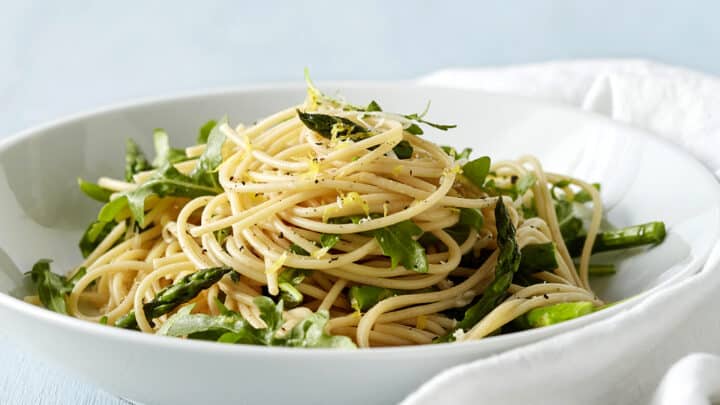 A white bowl of spaghetti mixed with green vegetables, garnished with herbs and lemon zest, sits on a light blue surface next to a white napkin—a fresh idea for June One Pot Dinners.