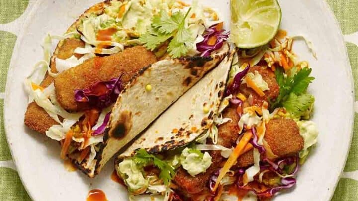 Two fish tacos on a white plate with shredded cabbage, carrot, cilantro, avocado, lime wedge, and hot sauce make for a healthy Mexican dinner atop a green and white polka dot tablecloth.