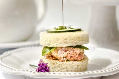 Elegant tea sandwich with a round cut, topped with cucumber and garnish, accompanied by a savory ham salad mix on a white plate with a delicate flower beside it. A teapot and more sandwiches rest gracefully on a cake stand in the blurred background.