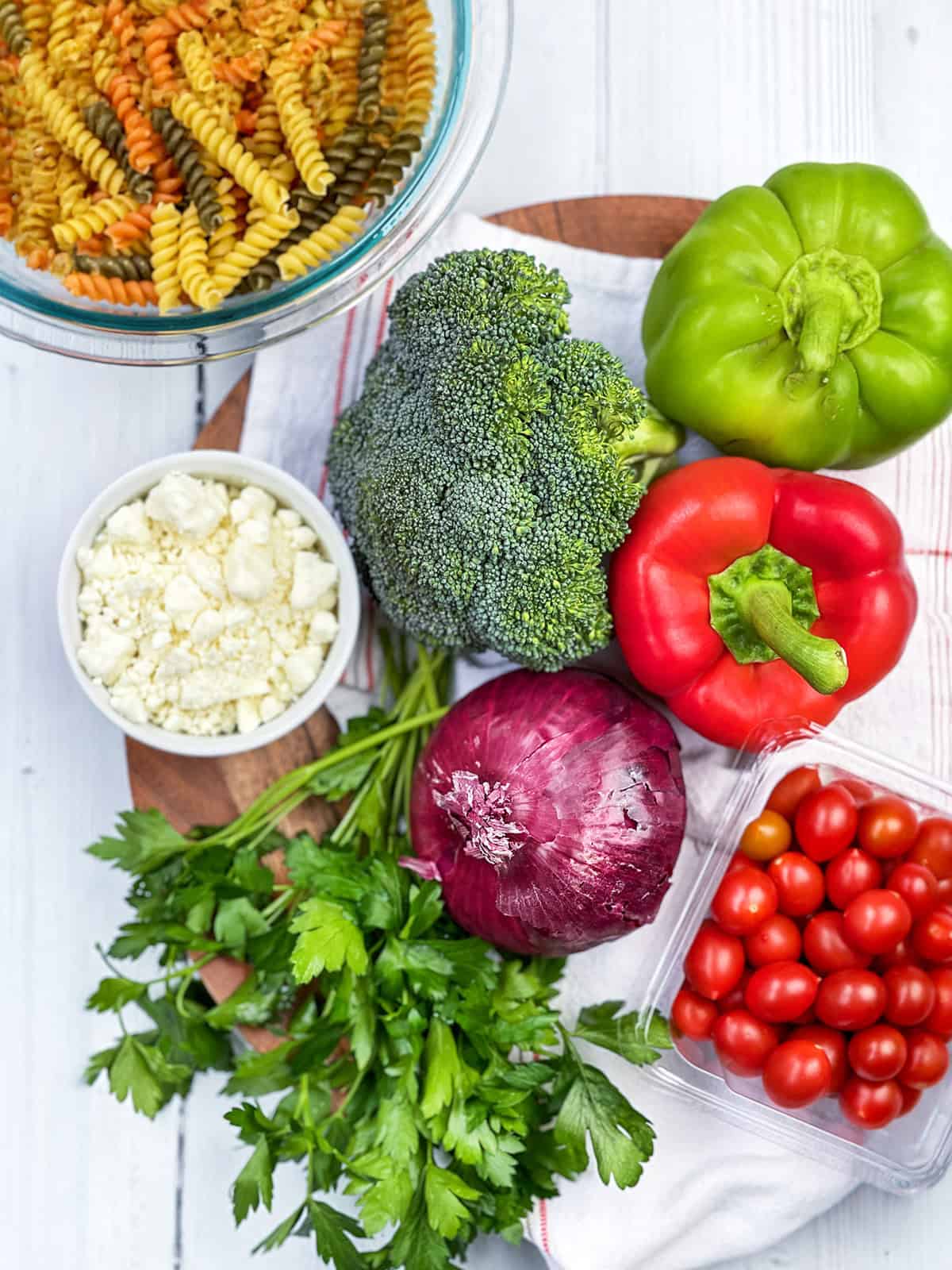 A bowl of tri-color rotini pasta, broccoli, green and red bell peppers, red onion, cherry tomatoes, parsley, and a small bowl of crumbled cheese arranged on a white surface creates the perfect summer pasta salad.