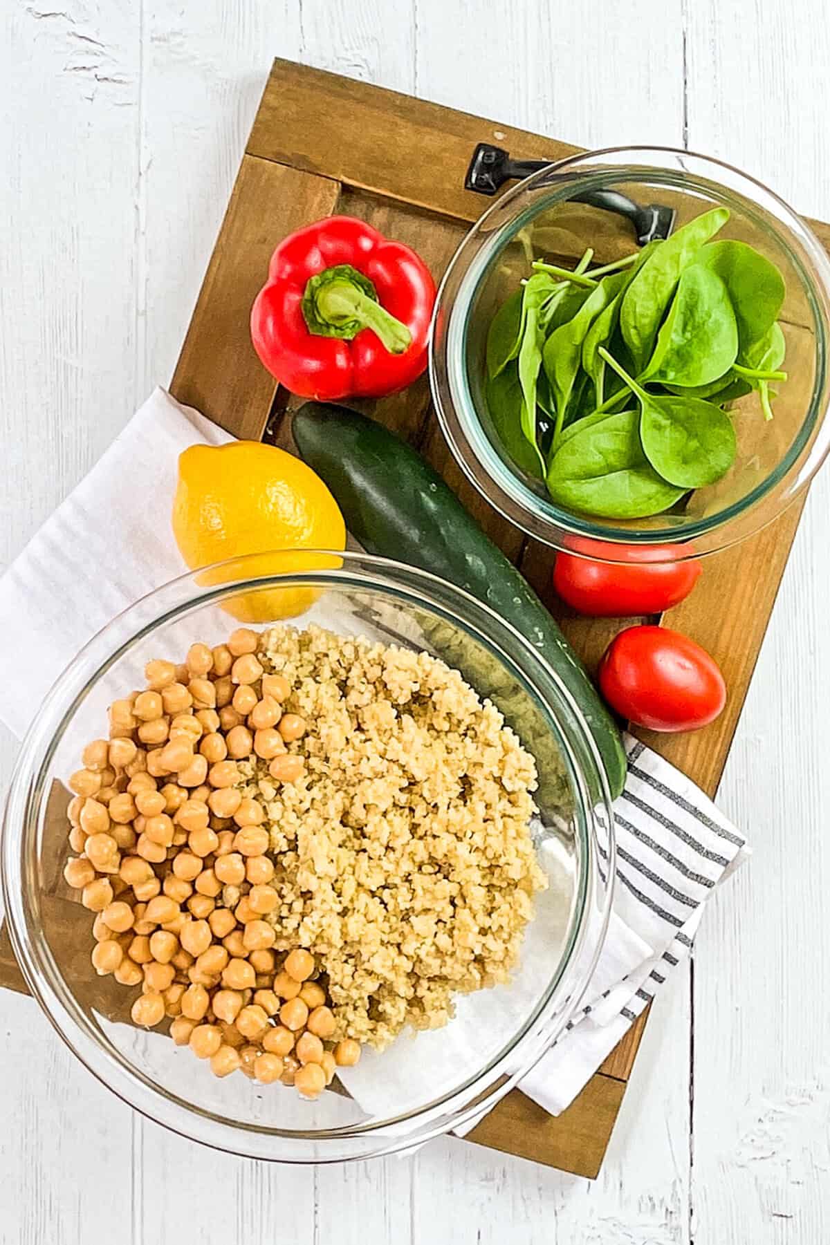 A wooden tray holds bowls of fresh spinach, chickpeas, and cooked quinoa, perfect ingredients for a vibrant quinoa salad, next to a red bell pepper, lemon, cucumber, and grape tomatoes on a white surface with a striped napkin.