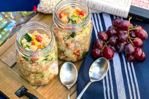 A picnic setup on grass with two jars of quinoa salad, a bunch of grapes, two spoons, a navy napkin, a book, sunglasses, and a blanket arranged on a wooden tray.