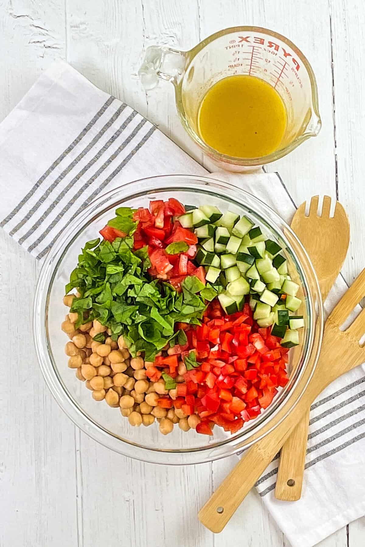 A glass bowl with chopped spinach, tomatoes, cucumber, red bell pepper, and chickpeas creates a colorful quinoa salad, set beside a measuring cup of dressing, wooden salad utensils, and a striped towel on a white wooden surface.