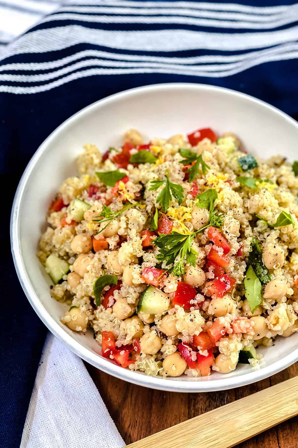 A white bowl filled with vibrant quinoa salad, featuring chickpeas, diced tomatoes, cucumber, red bell pepper, and fresh parsley, sits on a wooden surface with a blue and white striped cloth in the background.