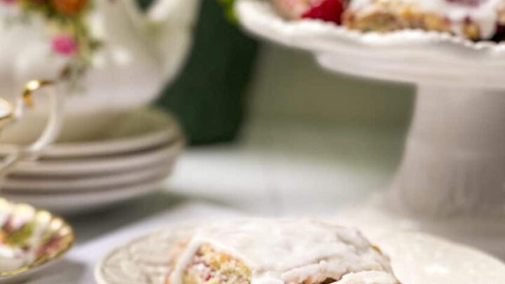 A raspberry scone with white icing sits on a plate, with more raspberry scones on a cake stand and a floral teapot in the background.