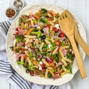 A large plate of colorful summer pasta salad with rotini, vegetables, olives, salami, and cheese is served with wooden utensils on a white cloth, accompanied by small bowls of spices nearby.