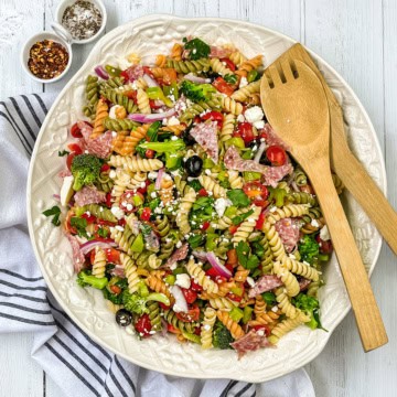 A large plate of colorful summer pasta salad with rotini, vegetables, olives, salami, and cheese is served with wooden utensils on a white cloth, accompanied by small bowls of spices nearby.