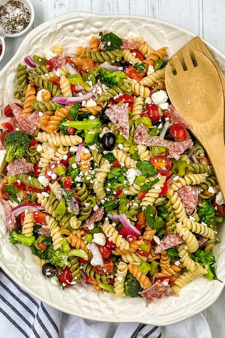 A large plate of colorful summer pasta salad with rotini, vegetables, olives, salami, and cheese is served with wooden utensils on a white cloth, accompanied by small bowls of spices nearby.