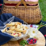 A picnic setup with a wicker basket, a plate of pinwheel pastries with berries, a small bowl of fresh berries, a white flower, and a dark cloth on a wooden surface.