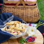 A picnic setup with a wicker basket, a plate of pinwheel pastries with berries, a small bowl of fresh berries, a white flower, and a dark cloth on a wooden surface.