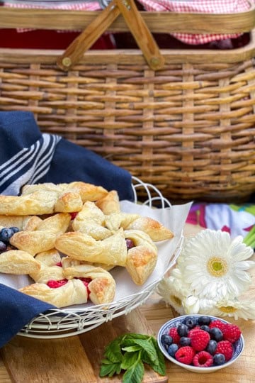 A picnic setup with a wicker basket, a plate of pinwheel pastries with berries, a small bowl of fresh berries, a white flower, and a dark cloth on a wooden surface.