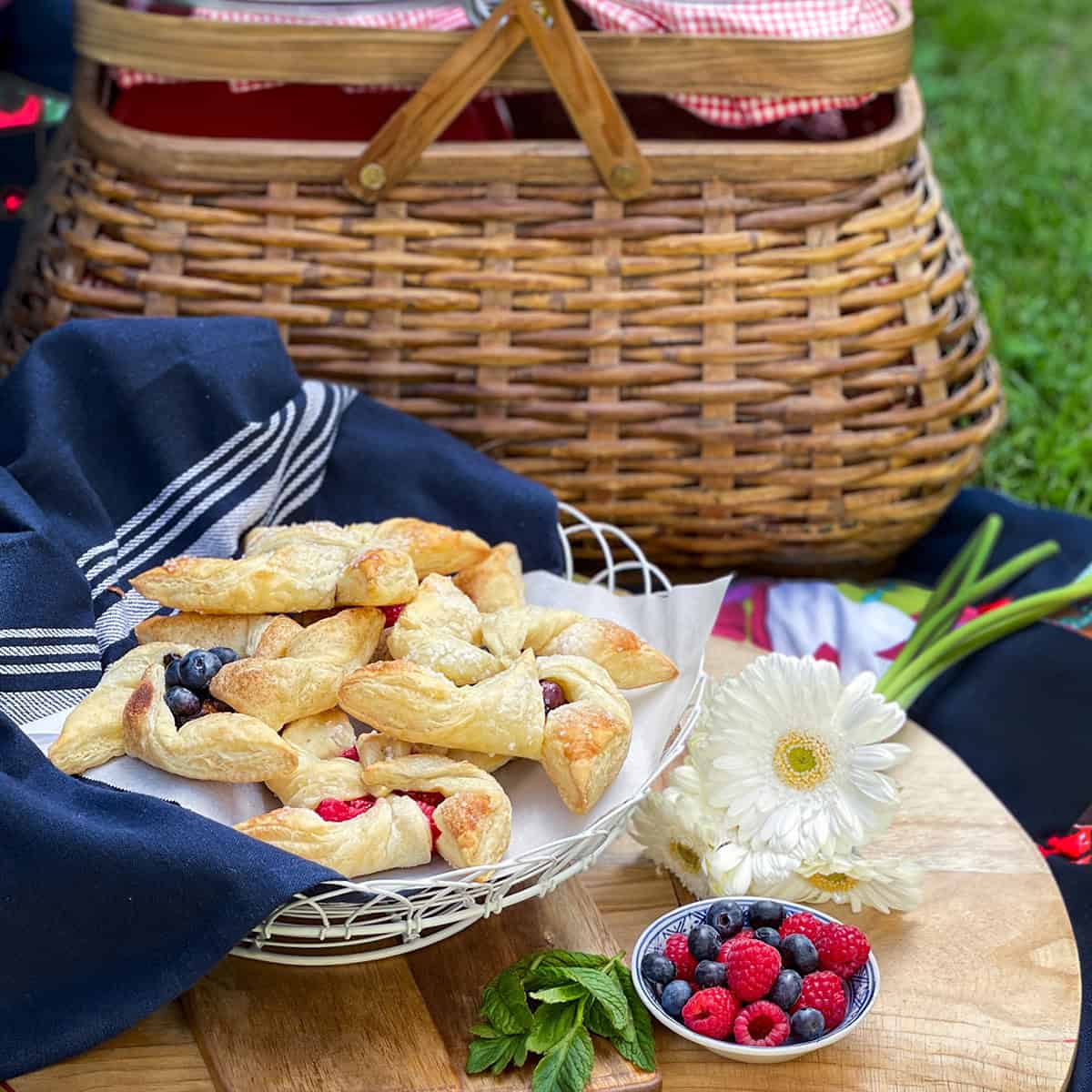 A picnic setup with a wicker basket, a plate of pinwheel pastries with berries, a small bowl of fresh berries, a white flower, and a dark cloth on a wooden surface.