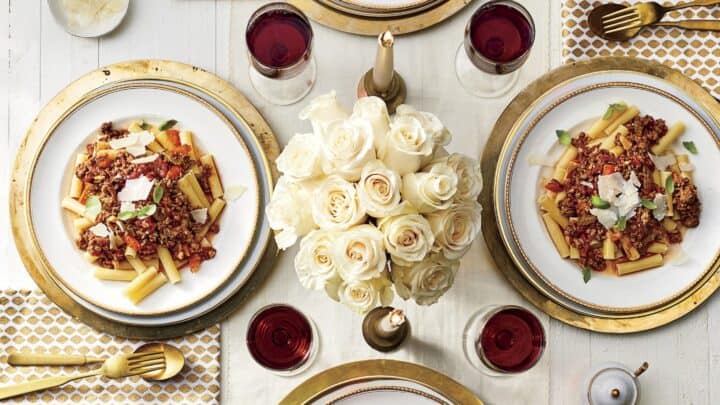 Four plates of pasta with meat sauce and cheese are arranged on a white table with gold-accented flatware, patterned napkins, red wine, and a centerpiece of white roses—perfect for Mother’s Day Dinner.