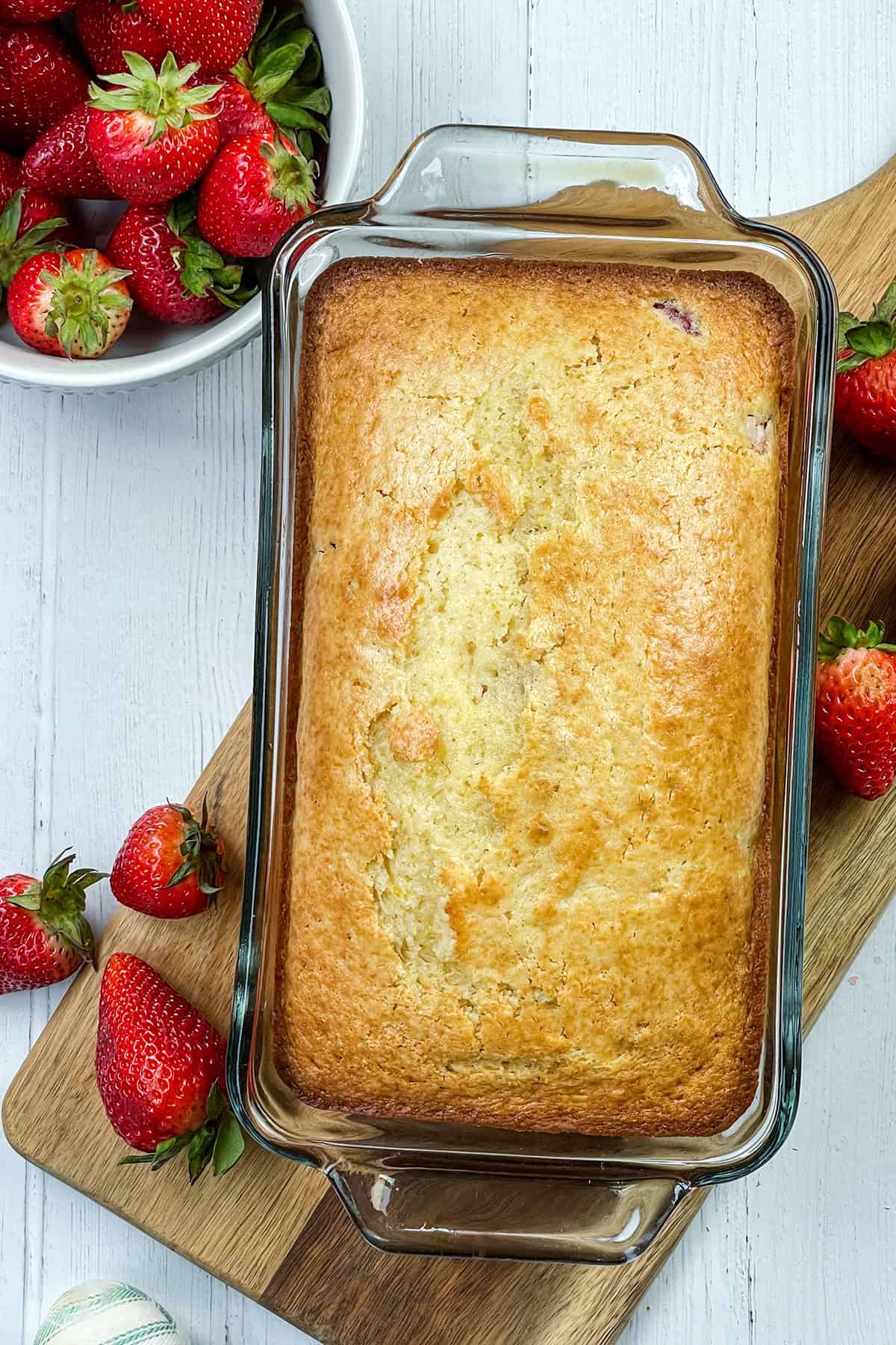 Glass baking dish with a golden-brown strawberry bread loaf on a wooden board, surrounded by fresh strawberries and a bowl of strawberries on a white table.