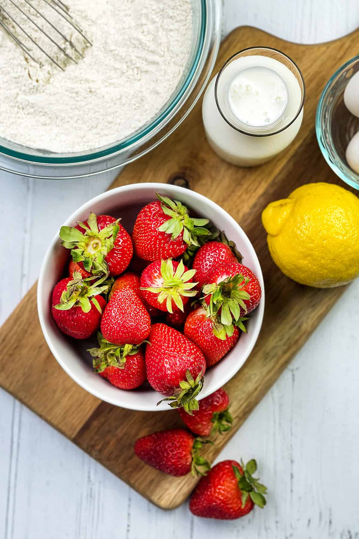 A bowl of fresh strawberries, a lemon, eggs, milk, and flour on a wooden board-everything you need to bake delicious strawberry bread, viewed from above.