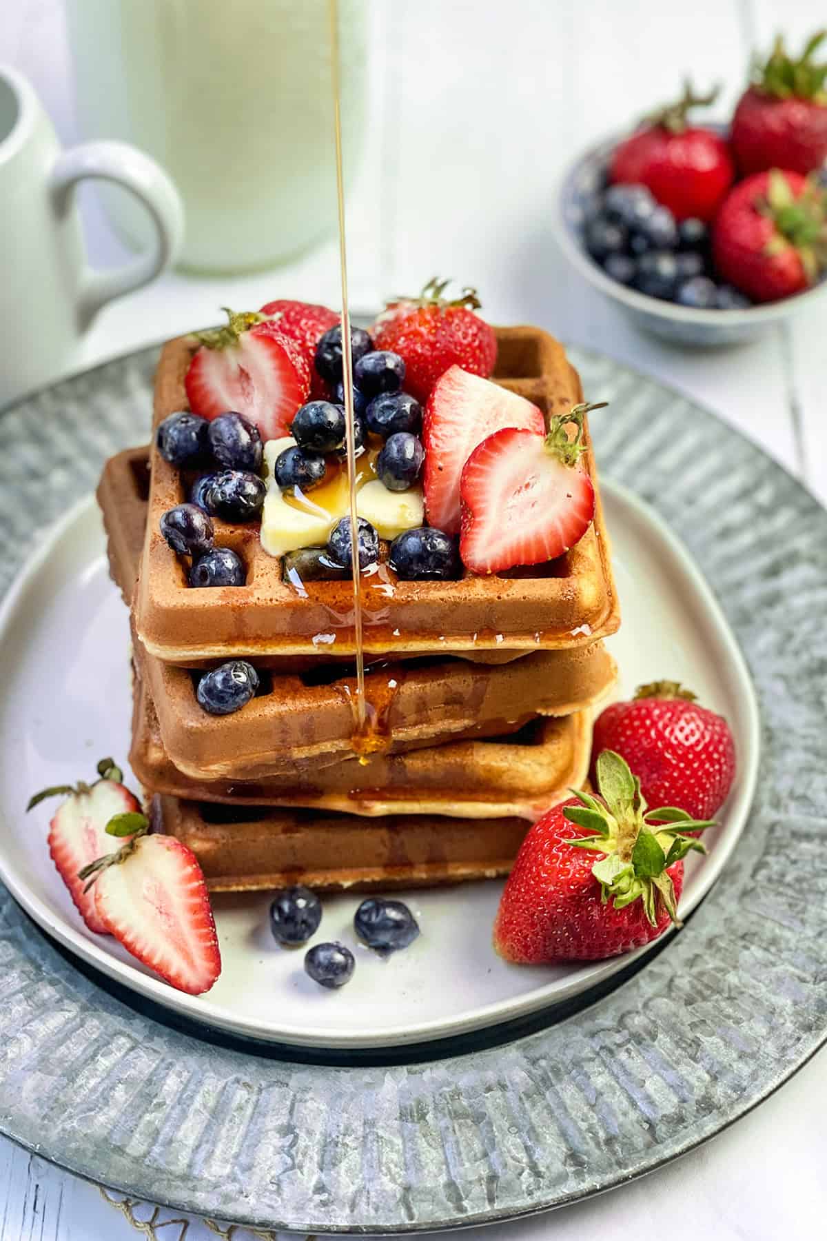 Stack of whole wheat waffles on a white plate with strawberries, blueberries, and a pat of butter with drizzling maple syrup.