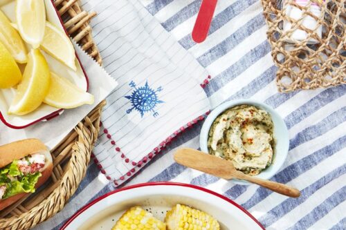 Plate of cut corn on the cob, lobster rolls with salad, lemon wedges, and herb butter on a striped tablecloth-perfect for trying out delicious summer picnic recipes.