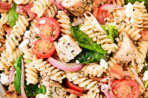 A bowl of rotini pasta salad with grilled chicken, cherry tomatoes, spinach, red onions, feta cheese, and herbs on a blue and white cloth-perfect for summer picnic recipes.