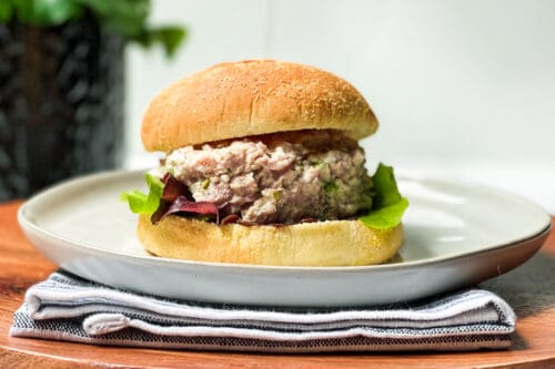 A ham salad and tuna salad sandwich with lettuce on a round bun sits on a white plate. The plate rests on a folded napkin atop a wooden surface. A blurred plant is in the background, adding a touch of green to the scene.