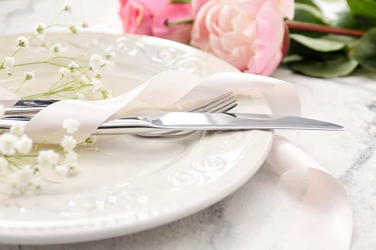 A white plate with silver cutlery wrapped in a pale pink ribbon and baby's breath flowers, next to pink roses on a marble surface&mdash;perfect for your elegant Mother's Day Dinner.