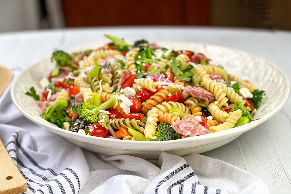 A bowl of summer pasta salad featuring rotini pasta, broccoli, cherry tomatoes, salami, feta cheese, and fresh herbs sits atop a white tablecloth.
