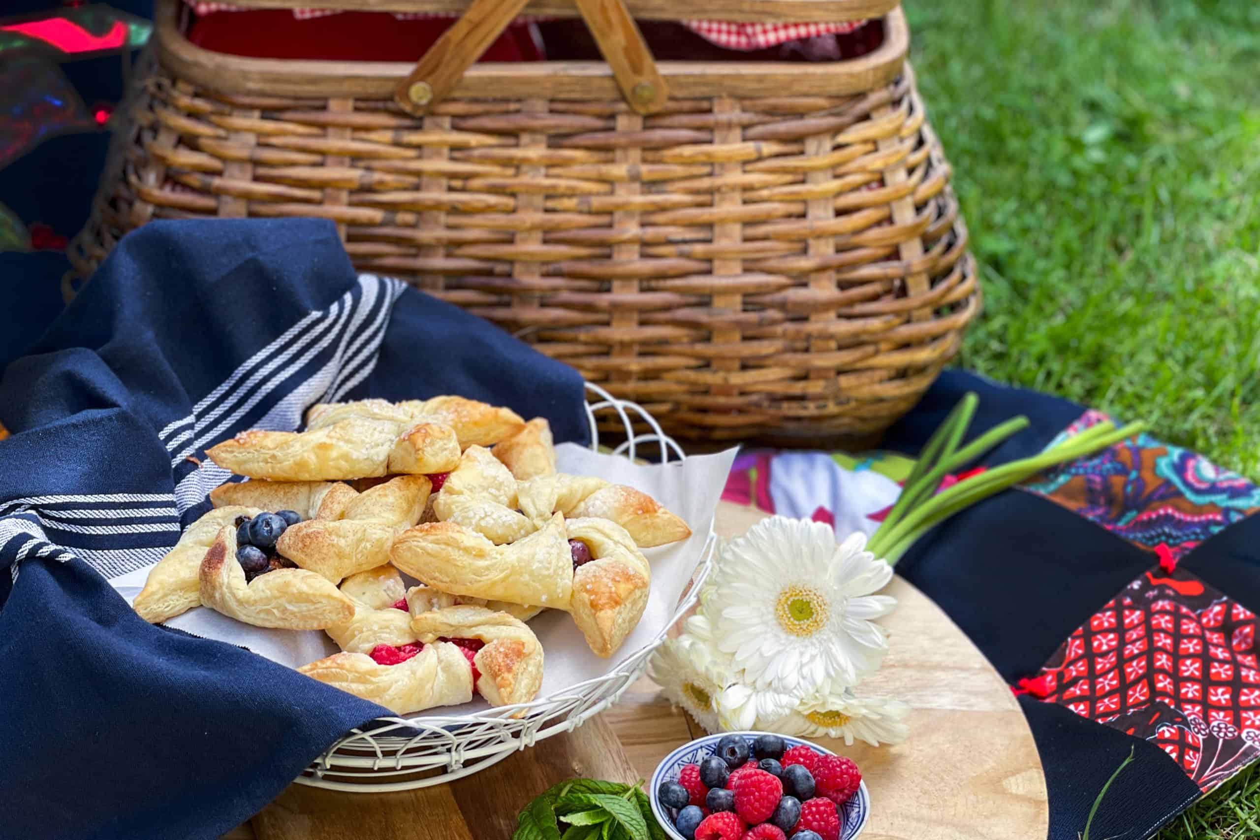 A picnic setup with a wicker basket, pinwheel pastries filled with fruit on a blue napkin, a small bowl of berries, and a white flower on a wooden board, all arranged on a blanket outdoors.