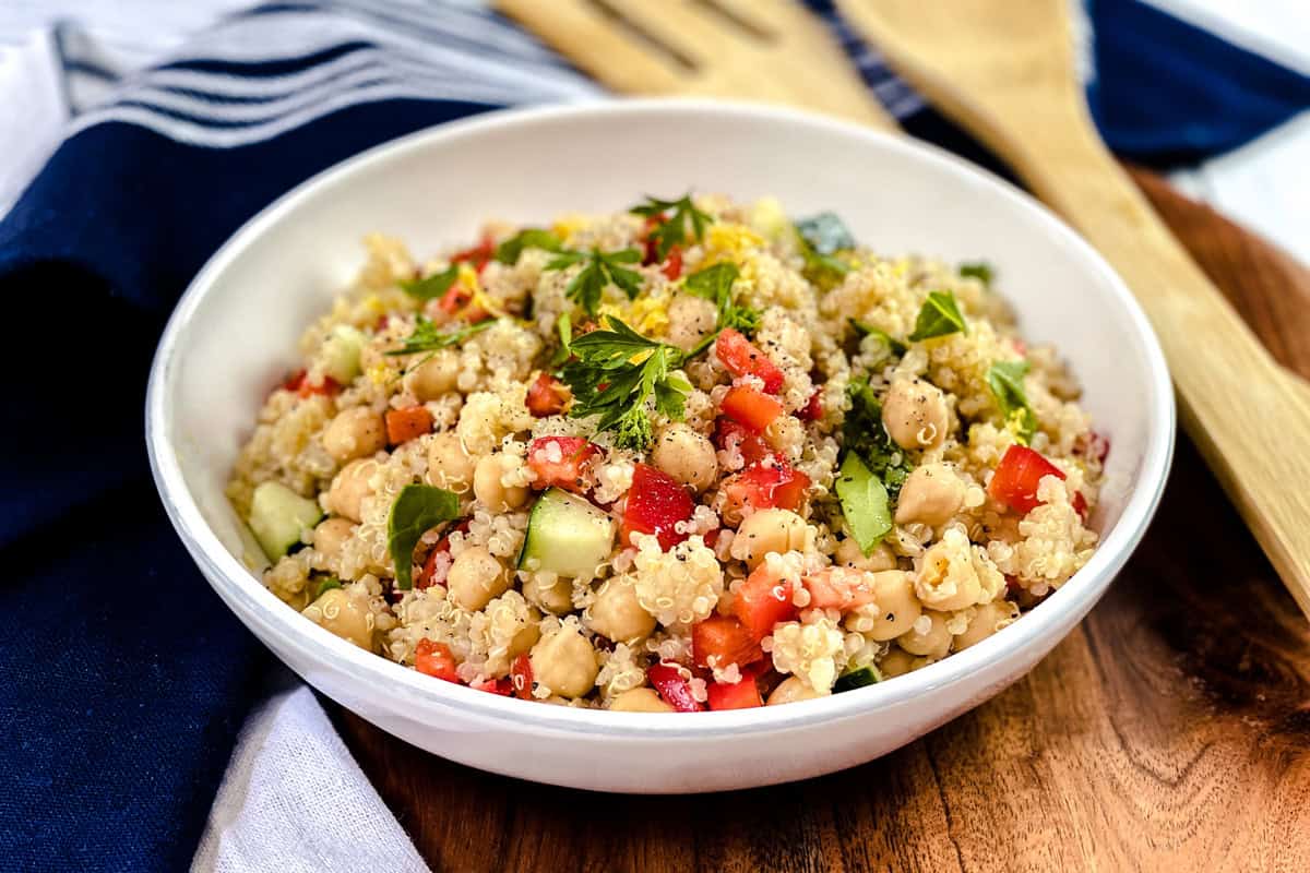A white bowl filled with quinoa salad, featuring chickpeas, diced tomatoes, cucumber, parsley, and herbs, sits on a wooden surface with a blue napkin and wooden utensils nearby. This vibrant quinoa salad is perfect for a fresh meal.