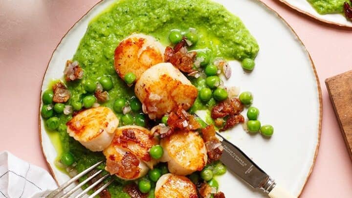 A plate of seared scallops with peas, bacon pieces, and green pea puree—perfect for Mother's Day Dinner—served with a fork and knife on a light pink table.