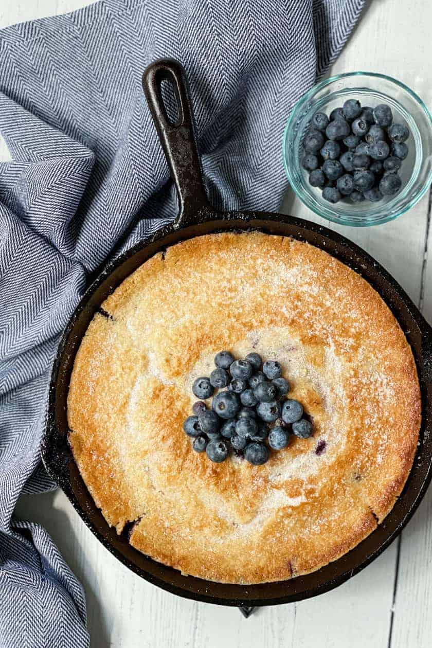 A skillet blueberry cobbler cake topped with fresh blueberries sits beside a small glass bowl of blueberries on a white wooden surface with a gray cloth.