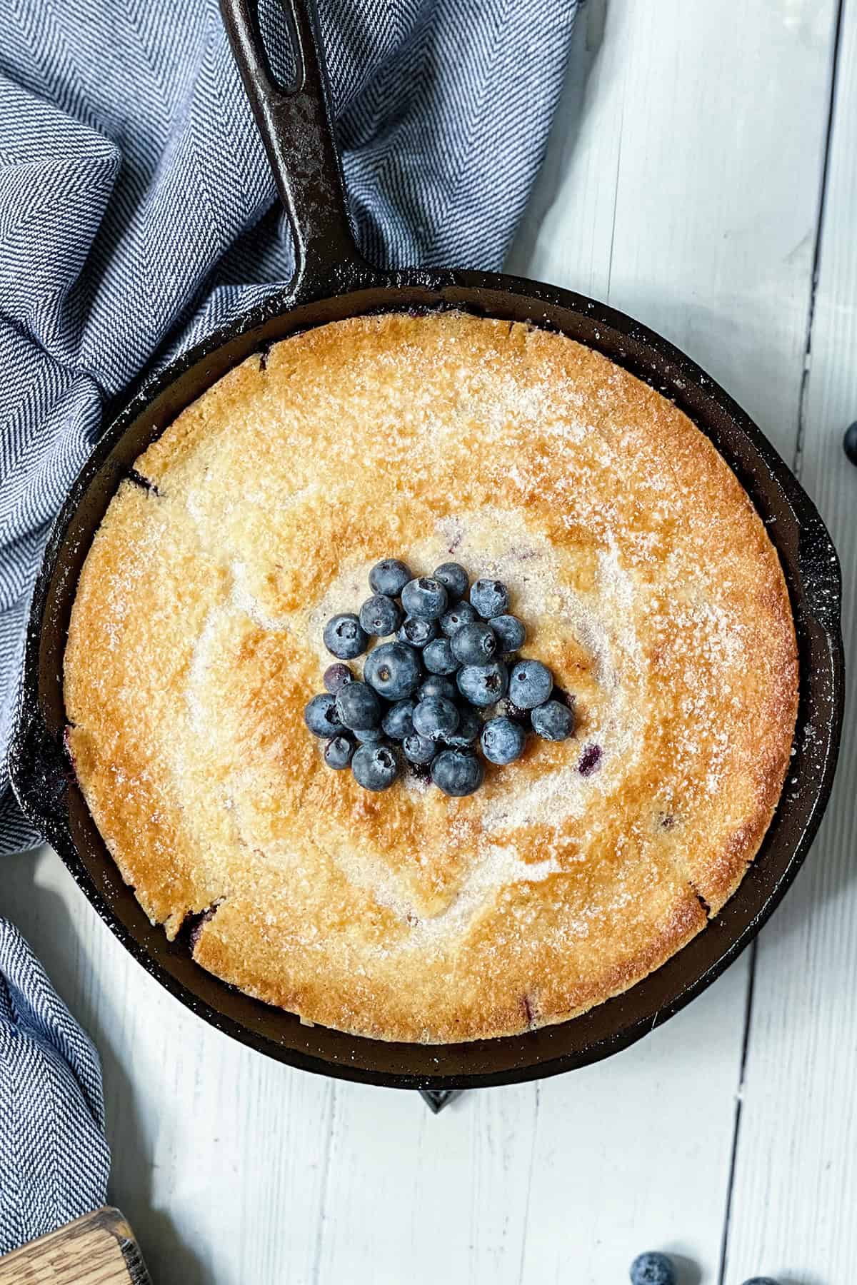 A golden-brown skillet blueberry cobbler topped with fresh blueberries sits on a light wooden surface next to a striped cloth.