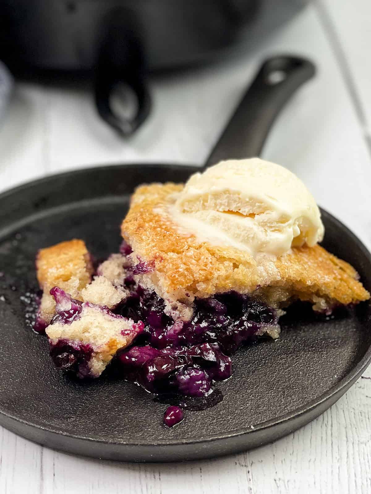 A serving of skillet blueberry cobbler topped with a scoop of vanilla ice cream on a black plate.