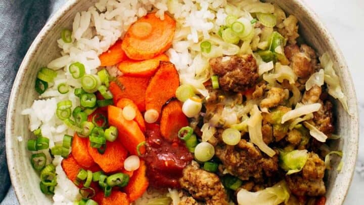 A bowl of white rice topped with sliced carrots, green onions, ground meat, cabbage, and a dollop of sauce—perfect for quick 15 Minute July Dinners—placed on a gray cloth.