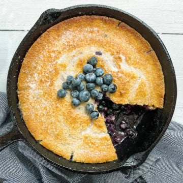 A golden-brown skillet blueberry cobbler in a cast iron pan, topped with fresh blueberries, with a slice removed to reveal the luscious filling.