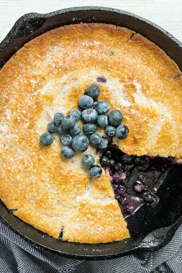 A golden-brown skillet blueberry cobbler in a cast iron pan, topped with fresh blueberries, with a slice removed to reveal the luscious filling.