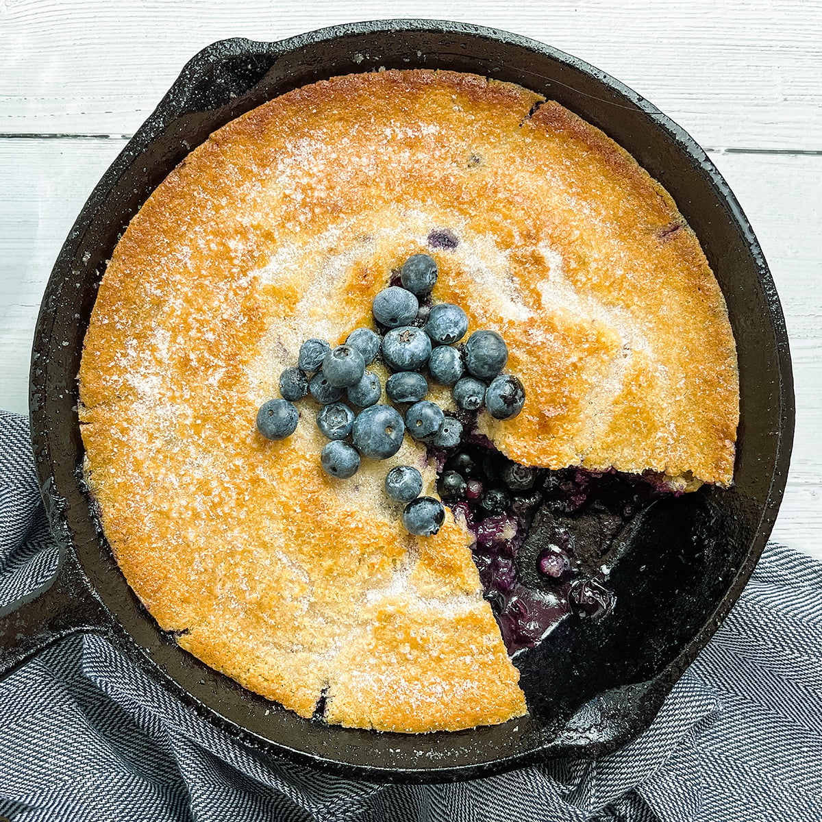 A golden-brown skillet blueberry cobbler in a cast iron pan, topped with fresh blueberries, with a slice removed to reveal the luscious filling.
