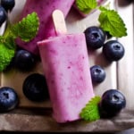 Blueberry popsicles rest on a tray, surrounded by fresh blueberries and green mint leaves for a colorful, refreshing display.