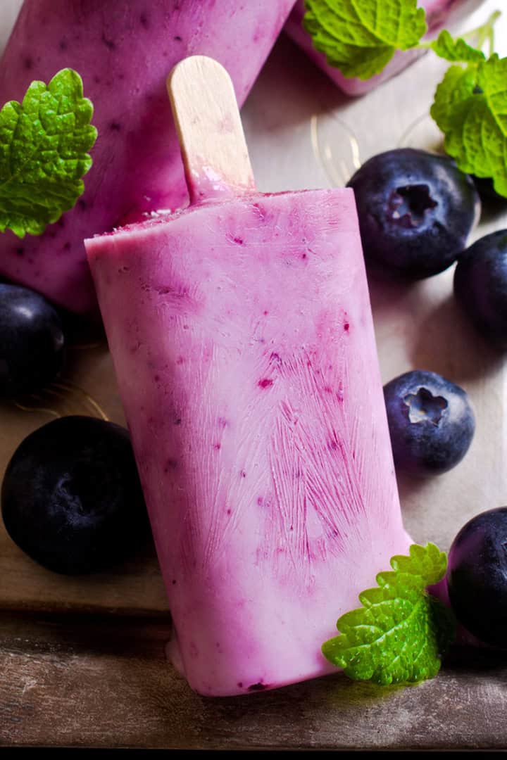 Blueberry popsicles rest on a tray, surrounded by fresh blueberries and green mint leaves for a colorful, refreshing display.