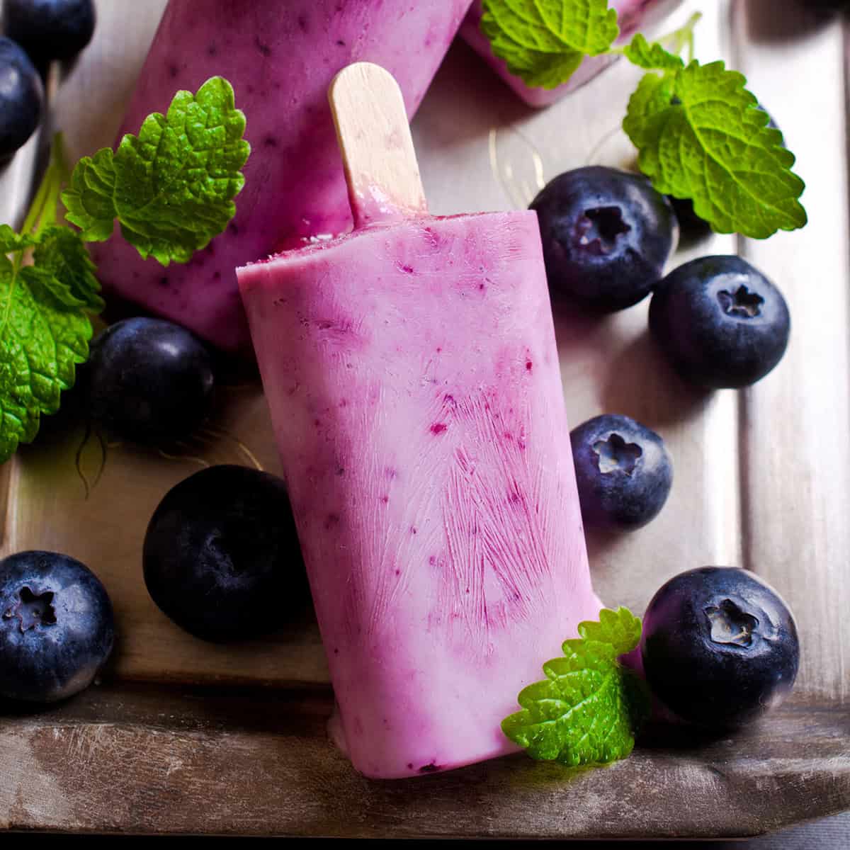 Blueberry popsicles rest on a tray, surrounded by fresh blueberries and green mint leaves for a colorful, refreshing display.
