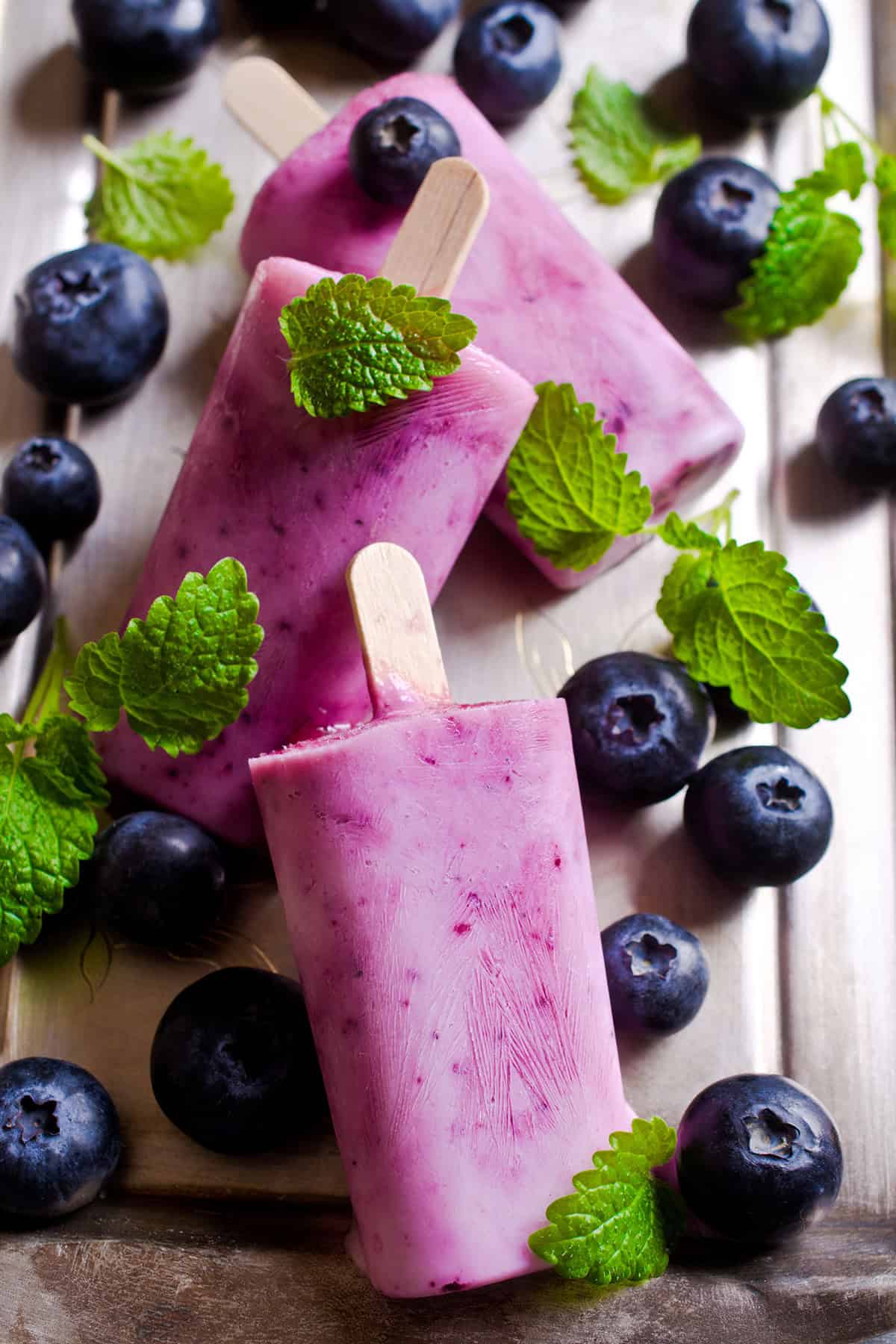 Three blueberry popsicles garnished with fresh mint leaves, surrounded by whole blueberries and mint on a metallic tray, create a refreshing summer treat.