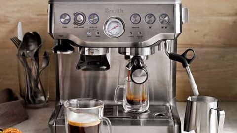 A stainless steel espresso machine brews coffee into a glass cup, with a mug of coffee, pastries, and milk frother nearby on a marble countertop—perfect inspiration for thoughtful Father's Day gifts.